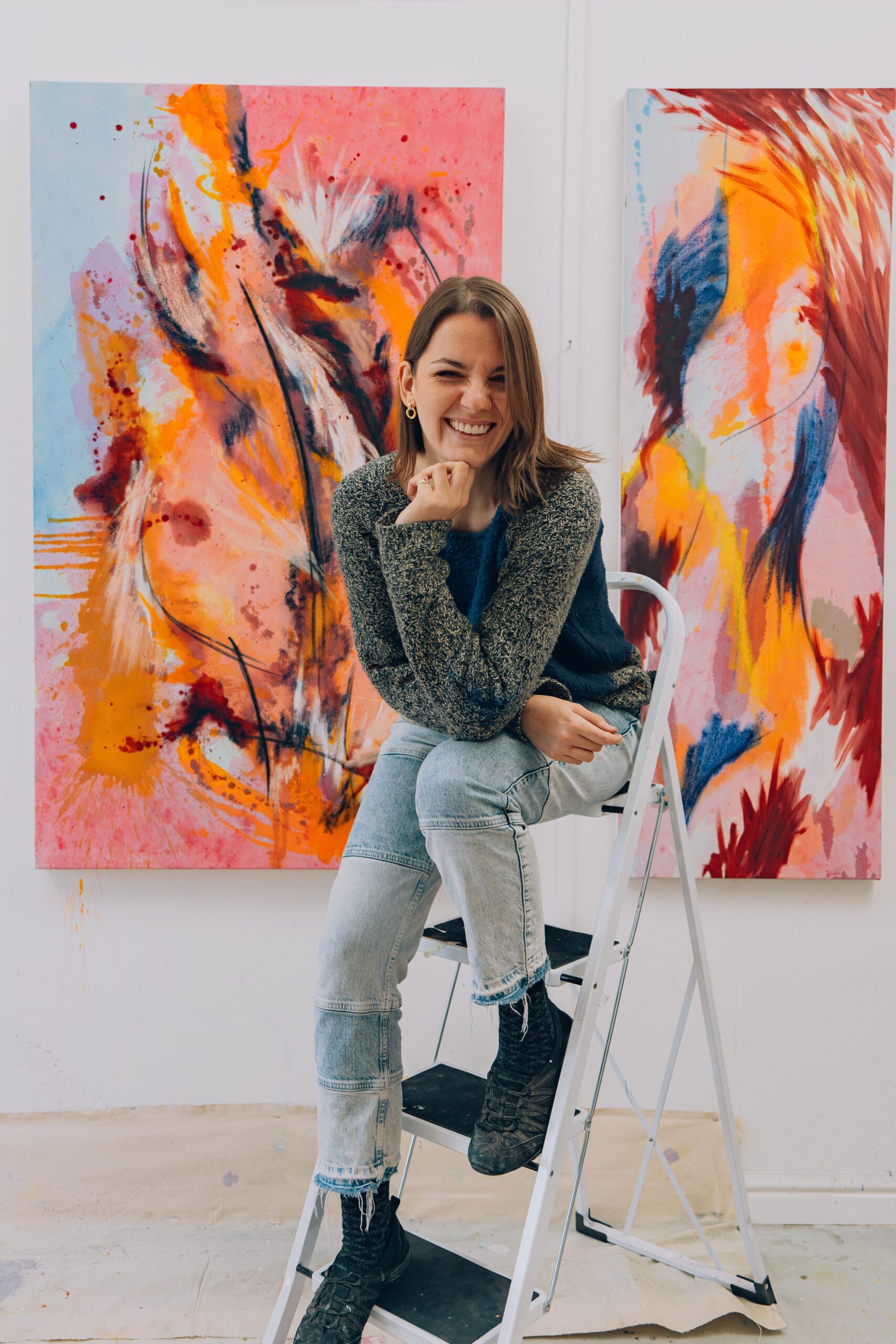 Portrait of artist Heather Green, sat on a stepladder in front of a bright pink, orange and blue painting. She holds her chin, rests her elbow on her knee, and appears to be laughing. 